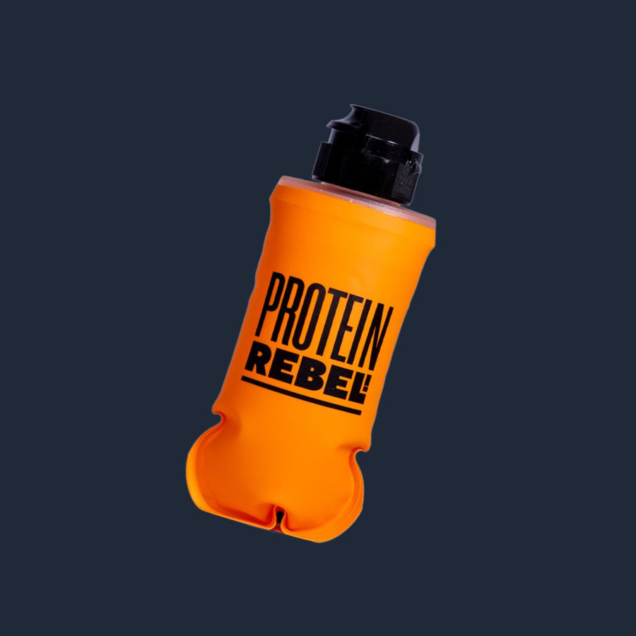 An orange collapsible gel flask with a black cap, featuring the brand name 'PROTEIN REBEL' printed in bold black letters on the front, set against a dark navy background.