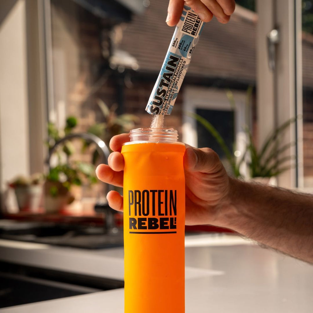 A hand holds an orange bottle labeled 'PROTEIN REBEL' while another hand pours powder from an electrolyte stick packet into the bottle, set against a blurred kitchen background.