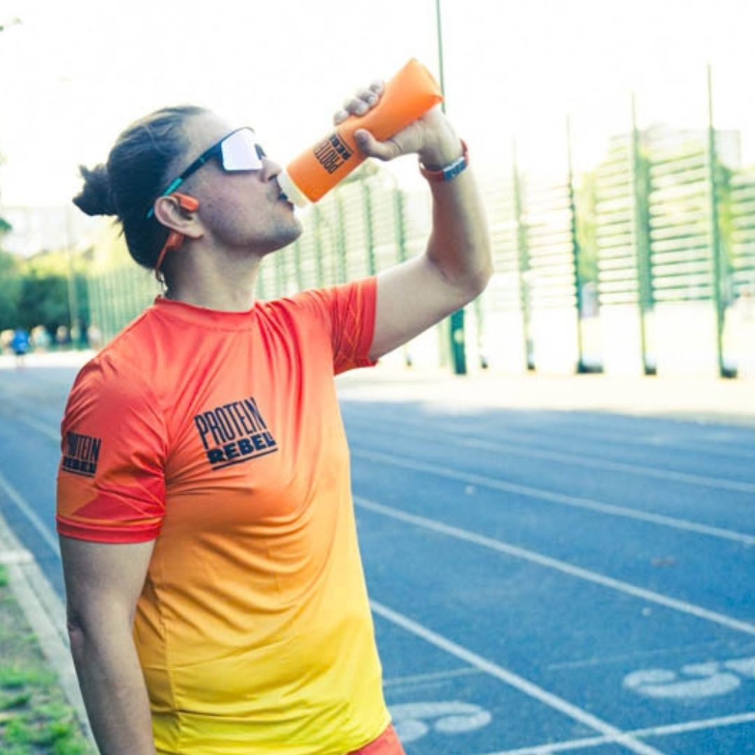 A man on an outdoor running track wearing a bright orange and yellow gradient athletic t-shirt with 'PROTEIN REBEL' printed on the chest, drinking from an orange bottle.