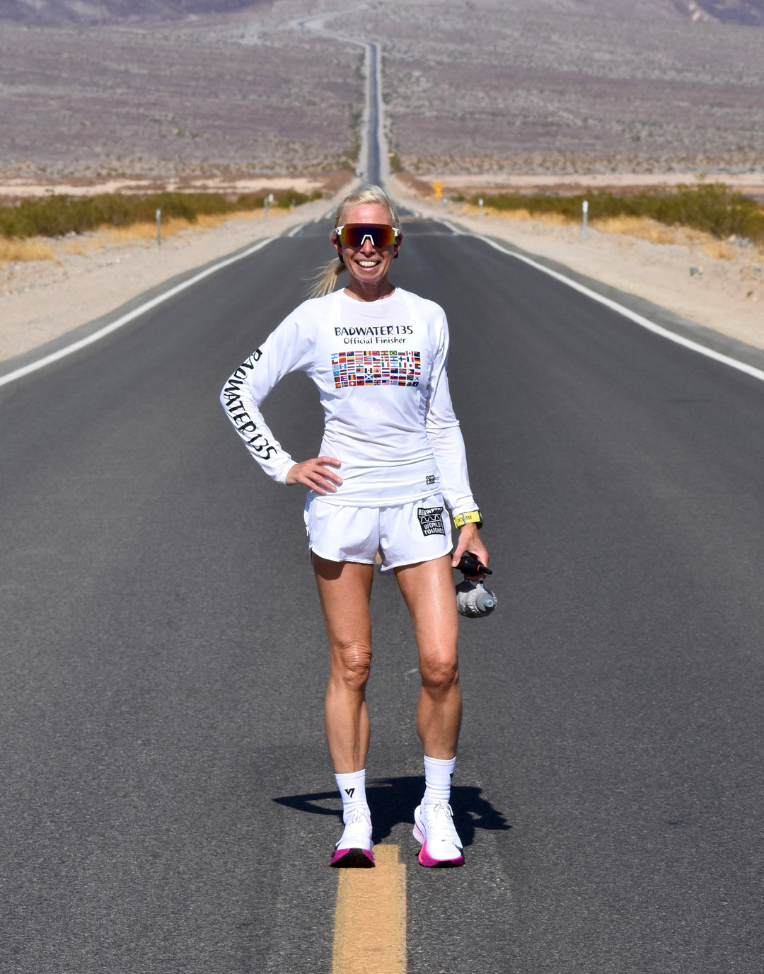 A woman stands on the center line of a desert highway wearing a white long-sleeve athletic shirt with 'UNABOLIZED' printed on the front and sleeve, white shorts, white socks, and pink running shoes.